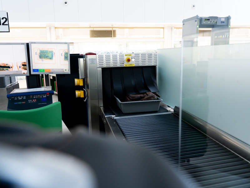 Wanne mit Gepäck wechselt von Röntgengerät auf Rollbahn (schwarze Rollen). Zugriffsschutz aus satiniertem Glas bzw. Milchglas sorgt für Sichtschutz